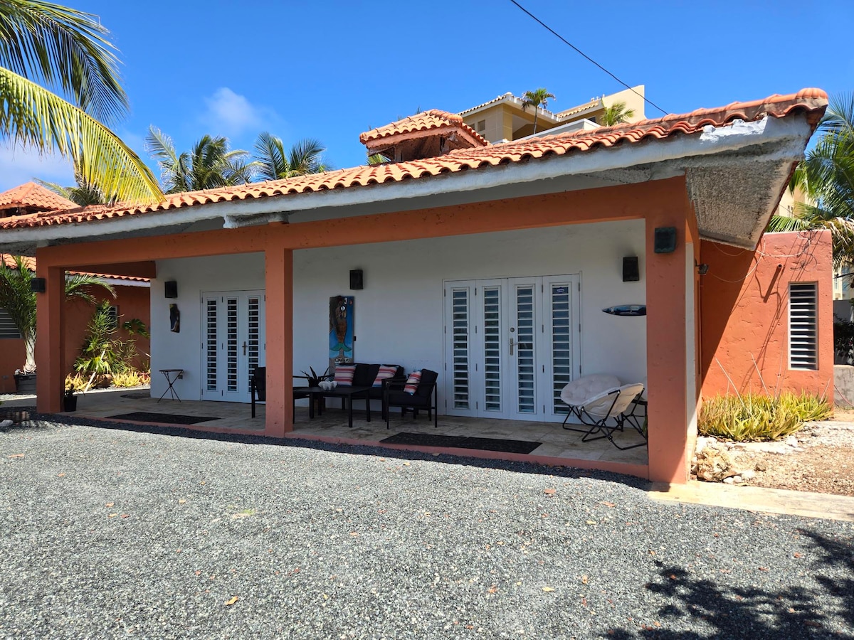 The exterior features a single-story bungalow with a textured orange facade and a tiled roof. Two sets of French doors open onto a comfortable porch area, furnished with lounge seating. Tropical plants surround the space, creating a relaxed atmosphere under a clear blue sky.