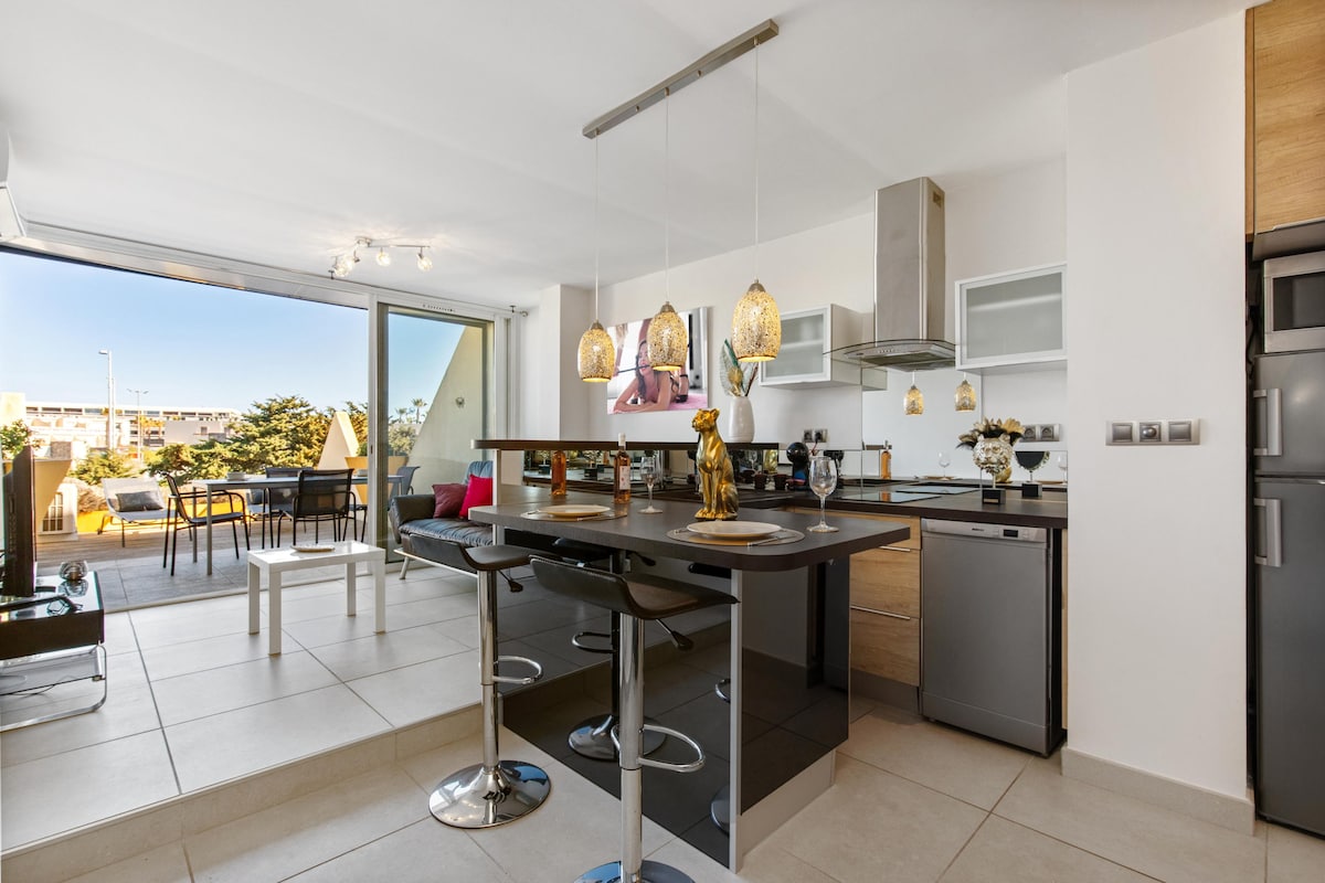 A modern kitchen space is observed, featuring a sleek island with high bar stools and contemporary appliances. Pendant lights offer soft illumination. A large glass door opens to a terrace with outdoor seating, providing an abundance of natural light and easy access to the outdoor area.