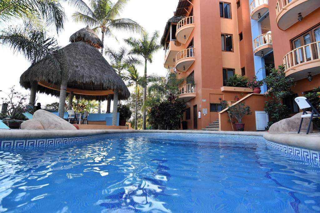 A sparkling outdoor pool reflects the surrounding palm trees. A thatched-roof gazebo provides shaded seating near the water's edge, while a multi-story building with balconies rises in the background, adorned with colorful flora.