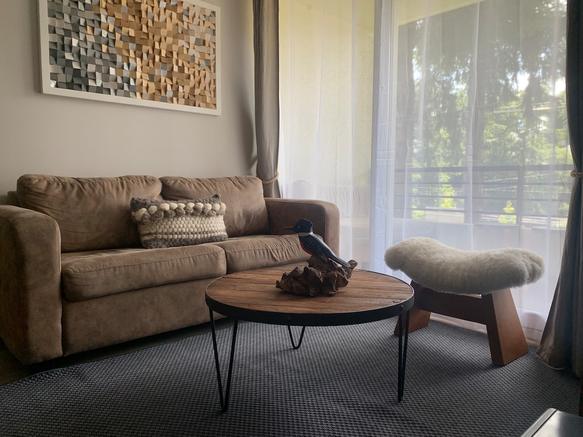 A comfortable living area features a soft brown couch adorned with decorative pillows. A round wooden coffee table sits at the center, with natural elements displayed on it. Sheer curtains allow diffused light to brighten the space, providing a view of the green surroundings.