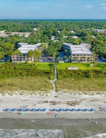 An aerial view presents a beachfront community with two buildings set among lush greenery. A sandy beach stretches in the foreground, dotted with blue umbrellas and beachgoers enjoying the ocean waves. Pathways lead from the buildings directly to the beach.
