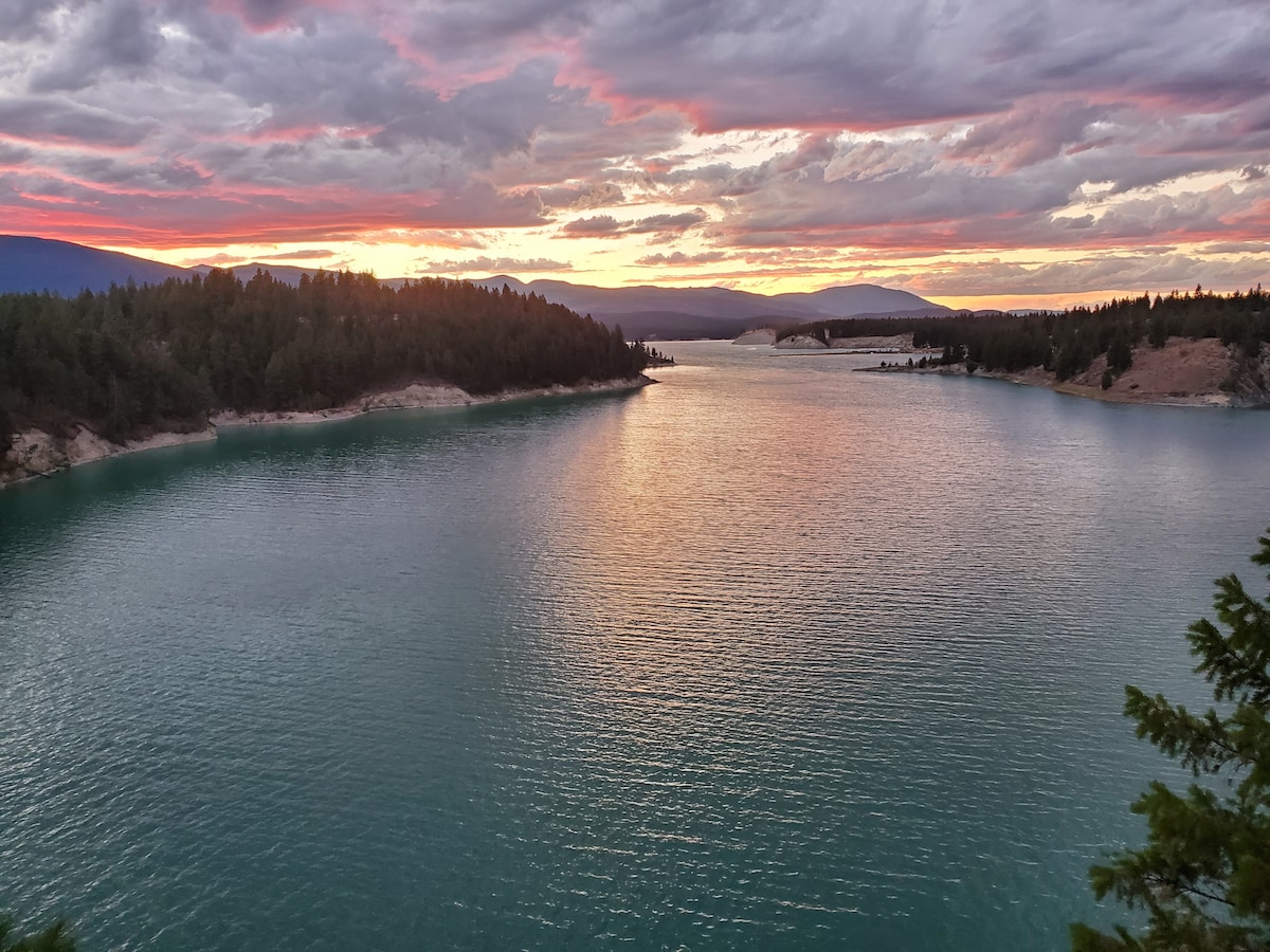A serene view of a lake at sunset is depicted, with gentle waves reflecting a palette of pink and orange hues across the water. Lush green trees line the shores, and distant mountains create a soft silhouette against the colorful sky.