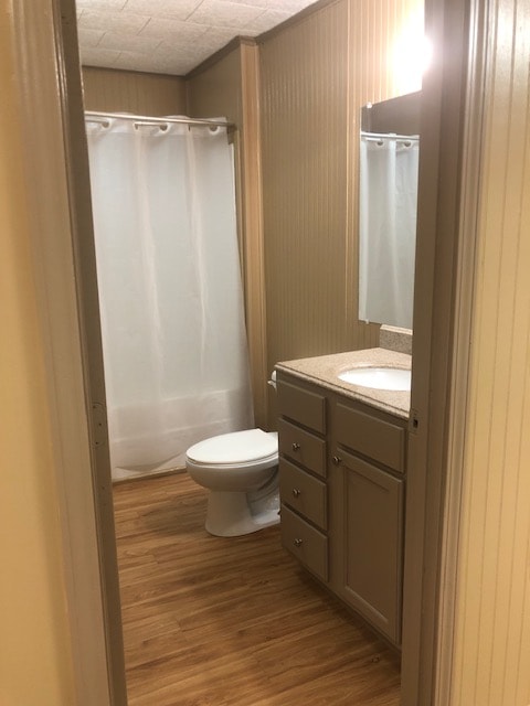A well-lit bathroom features a neutral color palette with tan wainscoting and wooden flooring. A shower with a white curtain is positioned alongside a toilet. A sink with a granite countertop and three drawers is reflected in the nearby mirror.