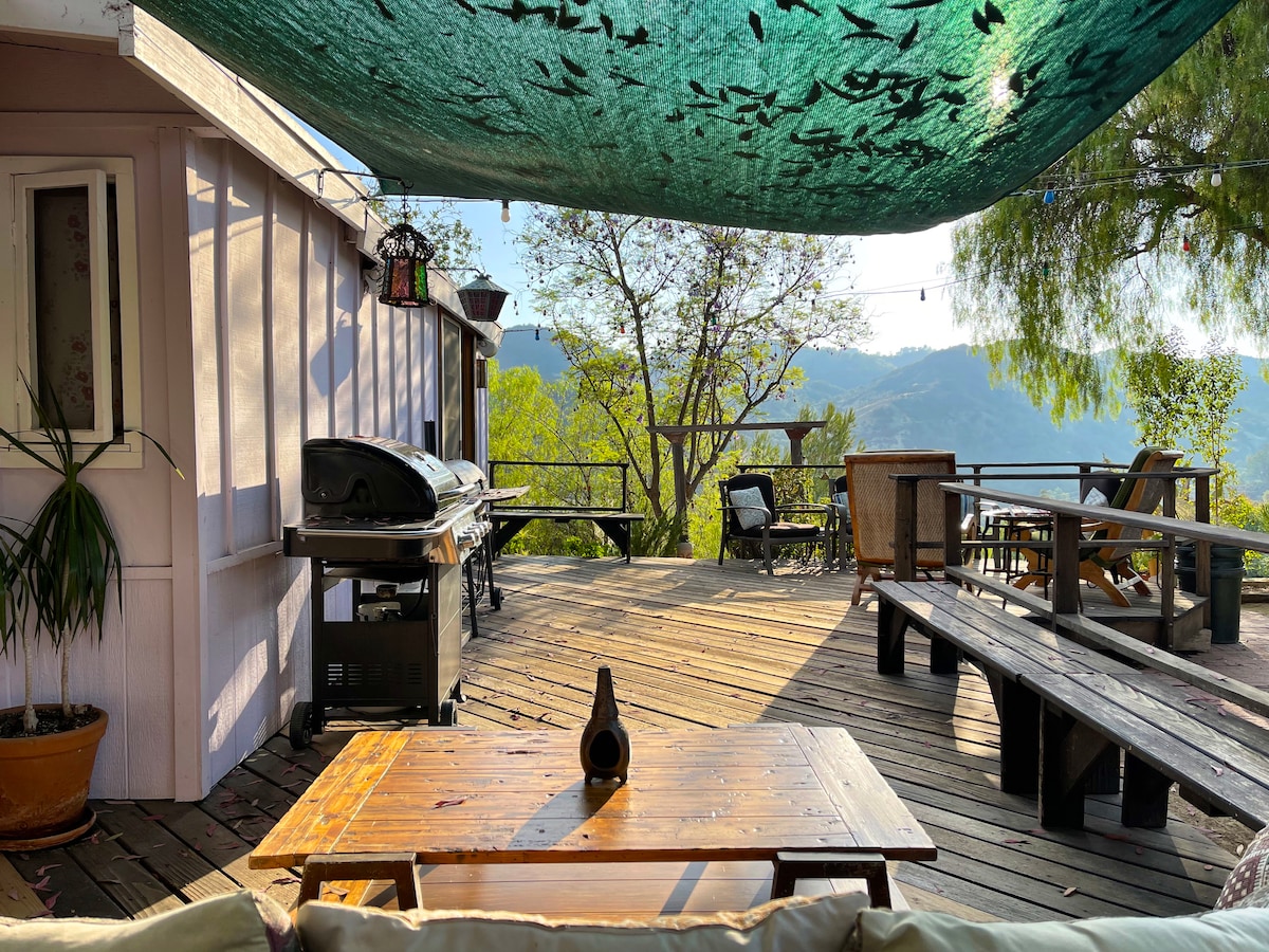 An outdoor deck is shown, featuring a wooden tabletop and a grill, surrounded by several seating arrangements. A canopy provides shade, while trees and mountain views are visible in the background. The space is designed for relaxation and socializing in a natural setting.
