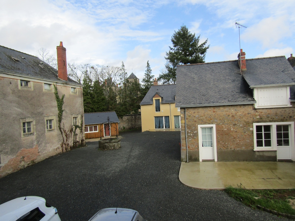 A view of a gravel courtyard is presented, surrounded by buildings with varied textures and colors. To the left, a weathered stone structure and a small cabin are visible. On the right, a modern building features large windows, while a tall tree and a tower are seen in the background.