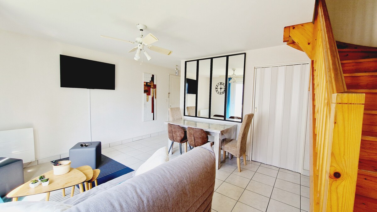 The living area features a comfortable sofa, a flat-screen television mounted on the wall, and a glass-top dining table accompanied by four chairs. A ceiling fan and large mirrors enhance the spacious feel. A staircase leads to the upper level, with white walls and tiled flooring throughout.
