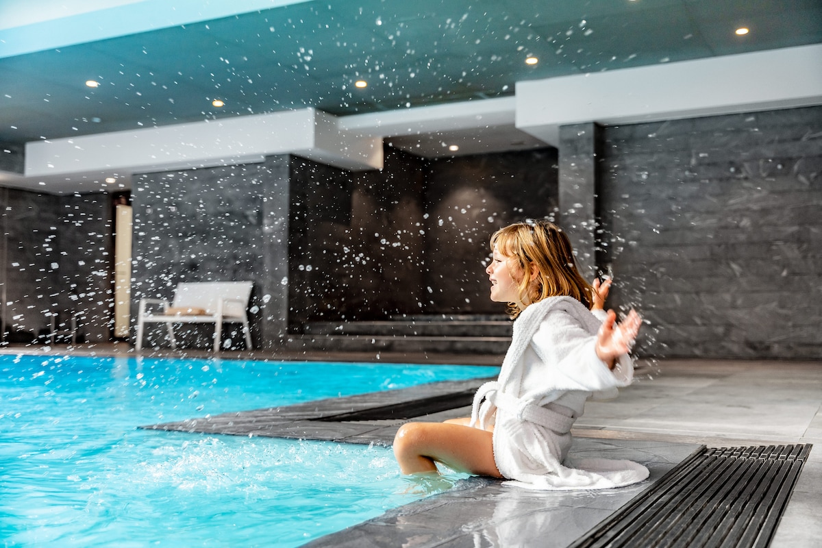 A child is seated at the edge of an indoor pool, water splashing around playfully. The pool features a clear blue surface, while the surrounding area is finished with sleek gray stone walls and well-lit by ceiling lights, enhancing the inviting environment.