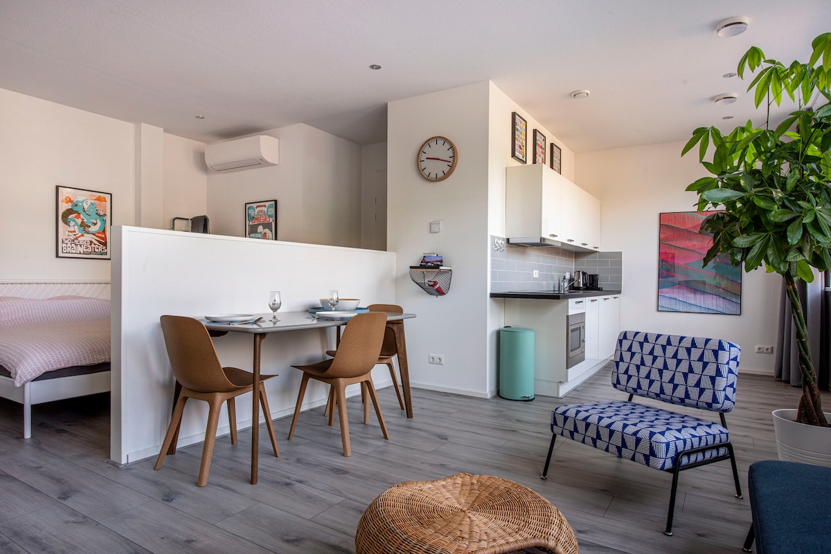 An open living space features a dining area with a table and chairs, adjacent to a modern kitchen equipped with essential appliances. A cozy seating area is highlighted by a patterned chair and a woven ottoman. Artwork decorates the walls, adding a touch of creativity.