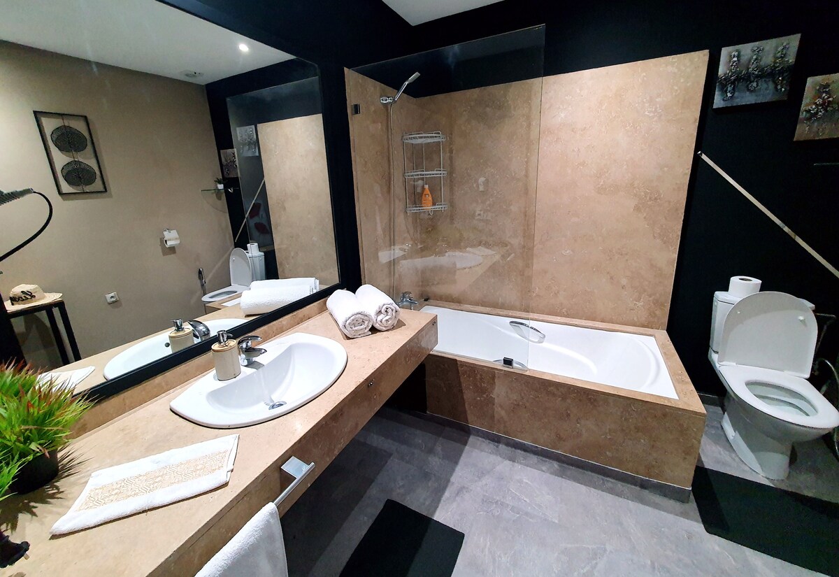 A modern bathroom is featured, displaying a sleek marble vanity with a large sink and stylish fixtures. A bathtub is positioned next to the shower area, surrounded by neutral-colored walls. Fresh towels are neatly arranged, and a minimalistic shelf holds toiletries.