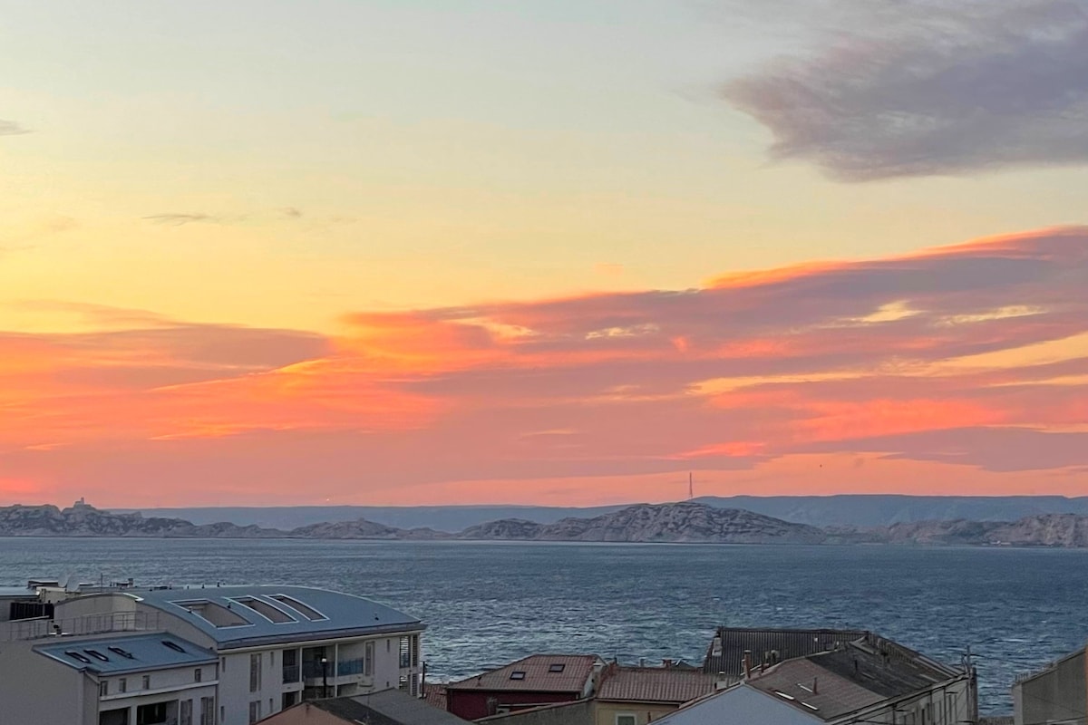 A serene seascape is captured during sunset, featuring vibrant hues of pink and orange reflecting on the water. The silhouette of the islands is visible in the distance, along with rooftops of nearby buildings in the foreground.