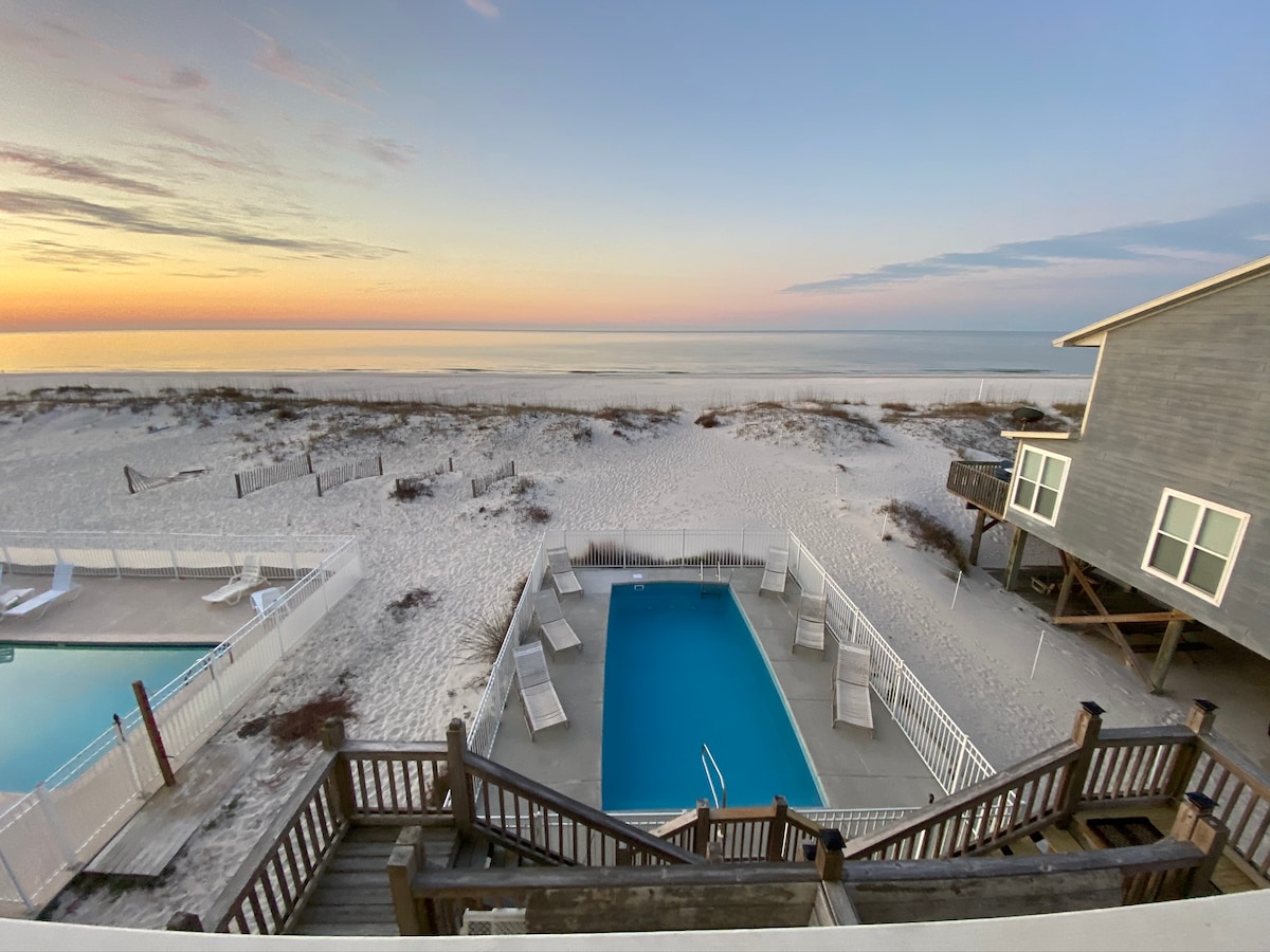 A view captures the beachfront heated swimming pool surrounded by lounge chairs, with soft sand leading to the tranquil gulf waters. The sunset fills the sky with gentle hues, while the duplex stands nearby, integrated with the coastal landscape.