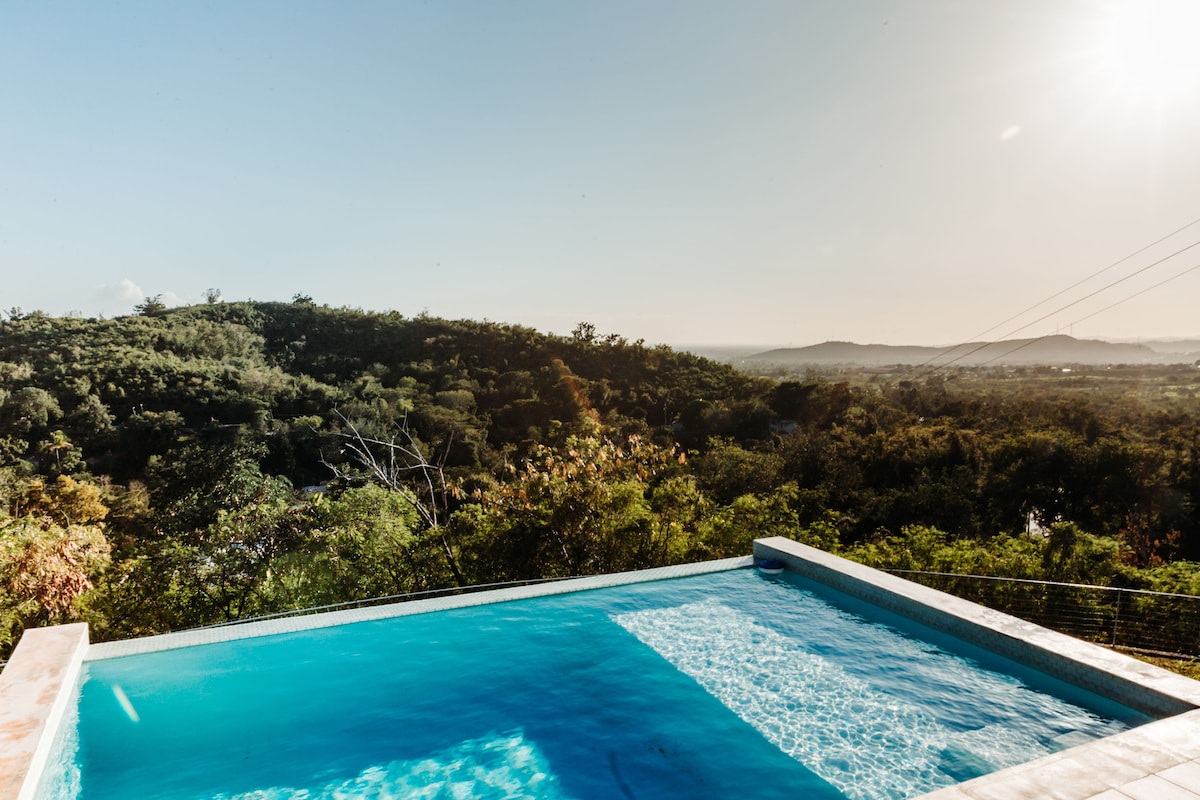 An infinity pool extends towards the landscape, creating a seamless transition between water and nature. Lush greenery surrounds the pool area, while distant hills are visible under a clear sky, illuminated by the sun. The tranquil setting invites relaxation and enjoyment of the views.