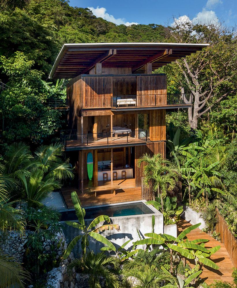 The modern treehouse showcases a three-story structure crafted from natural wood, surrounded by lush greenery. A small pool is visible at the base, with spacious outdoor areas leading up to the open air king suite on the top level, punctuated by large windows allowing for natural light.