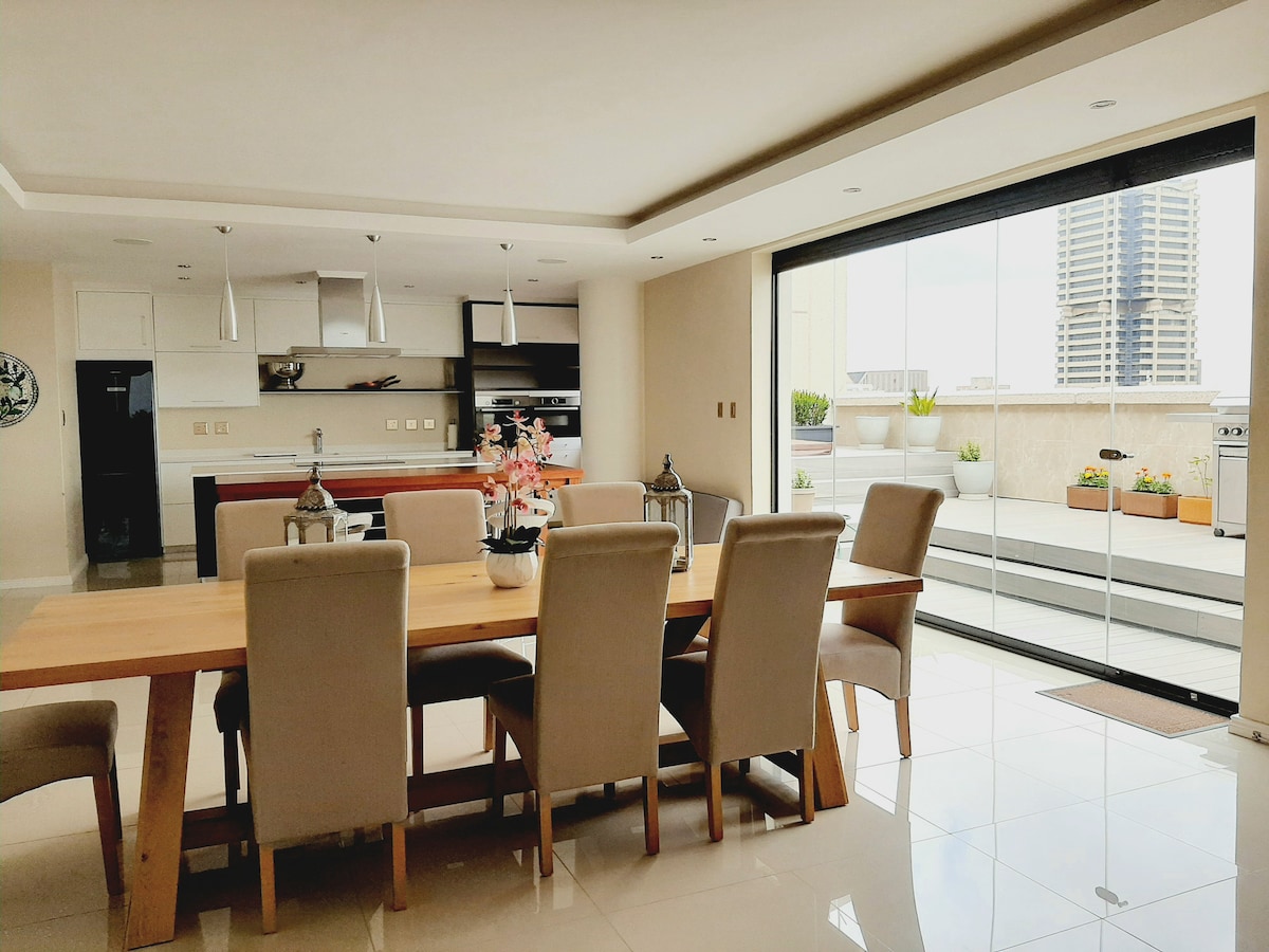 An open-plan dining area features a long wooden table surrounded by upholstered chairs. Large sliding glass doors invite natural light and provide views of an outdoor terrace, while a modern kitchen is visible in the background, equipped with sleek cabinetry and contemporary appliances.