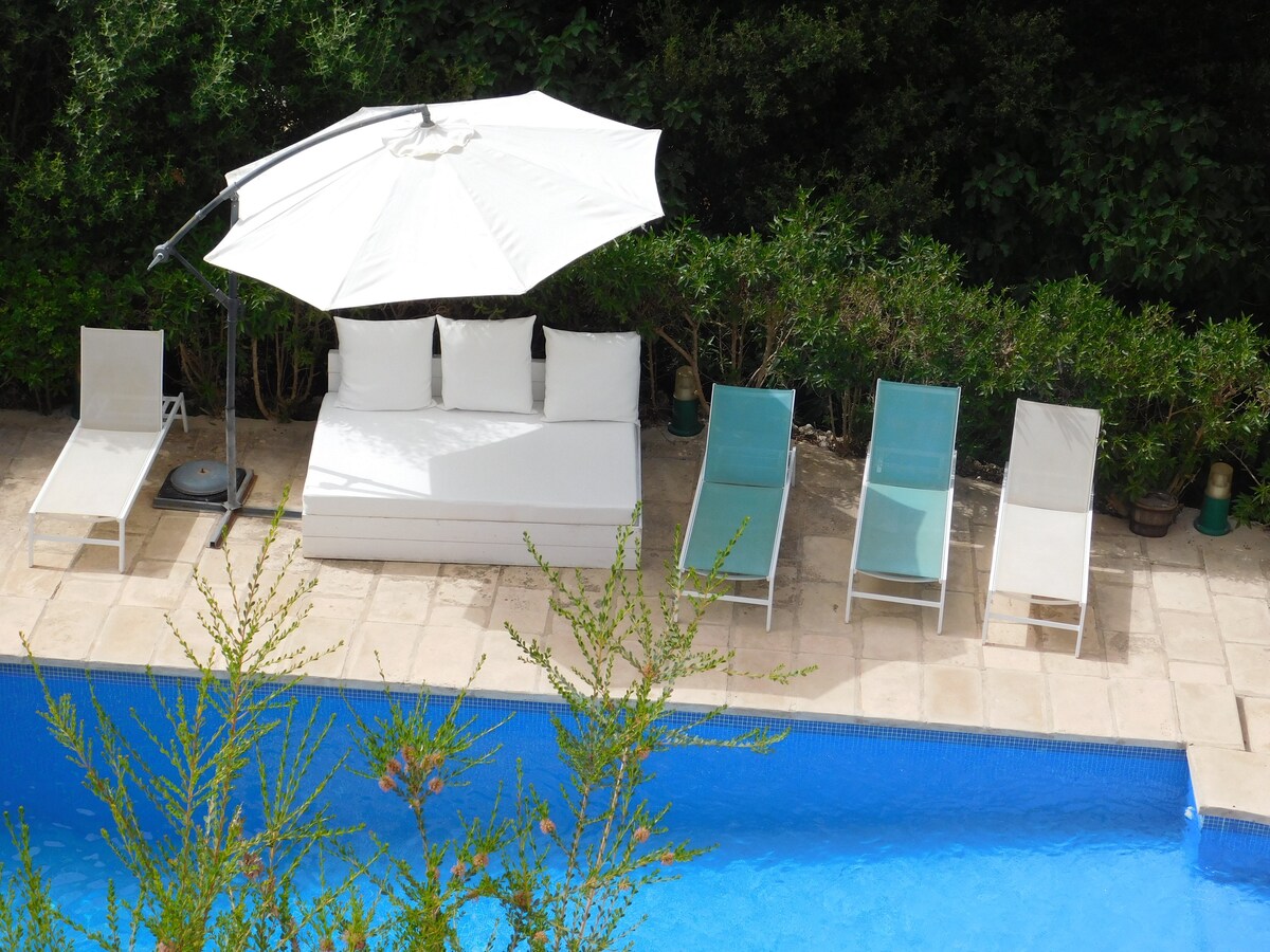 A clear view of a swimming pool surrounded by a sun terrace. A large white umbrella shades a cushioned seating area, while four lounge chairs are arranged nearby. Lush greenery frames the area, providing privacy and tranquility.