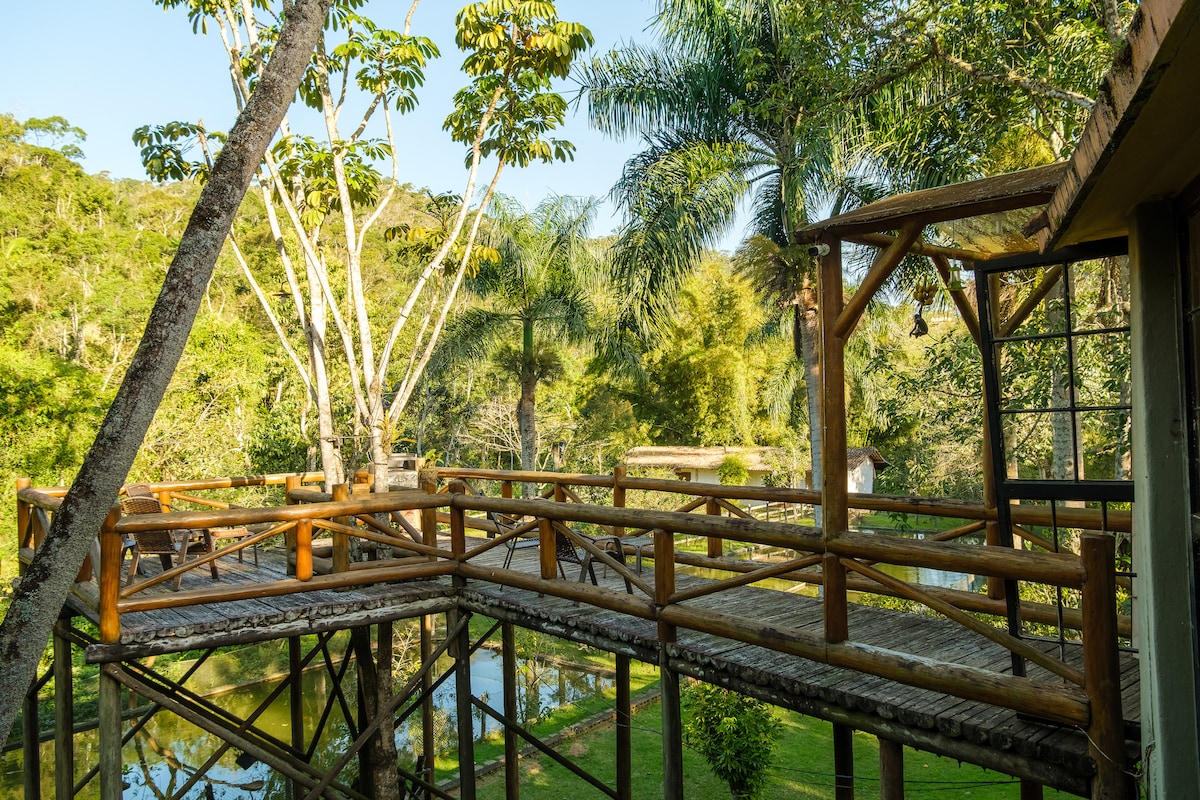 A wooden deck elevated above the ground is framed by lush greenery and tall trees. The surrounding landscape includes a tranquil pond reflecting the natural beauty, creating a serene atmosphere. The structure is designed to blend harmoniously with the forest environment.