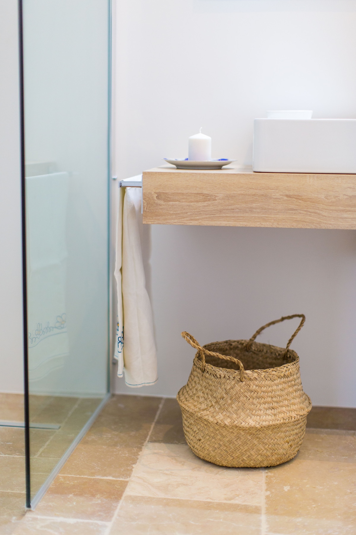 A clean bathroom space features a glass shower stall and a wooden floating vanity. A white candle is placed on the counter next to a small stack of toiletries. A woven basket is positioned on the floor, adding a touch of natural texture to the design.
