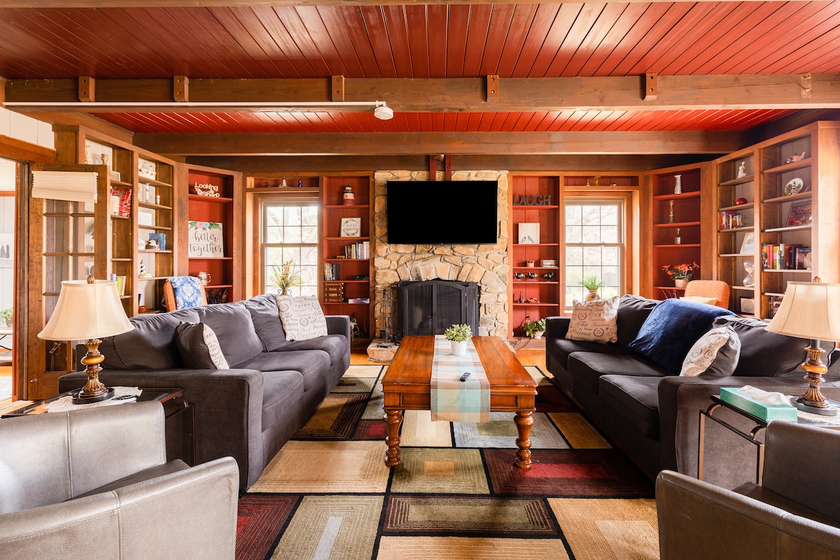 A spacious living area features comfortable seating on dark sofas arranged around a central wooden coffee table. A stone fireplace serves as a focal point against a backdrop of shelves filled with decor and books. Large windows allow natural light to fill the room.