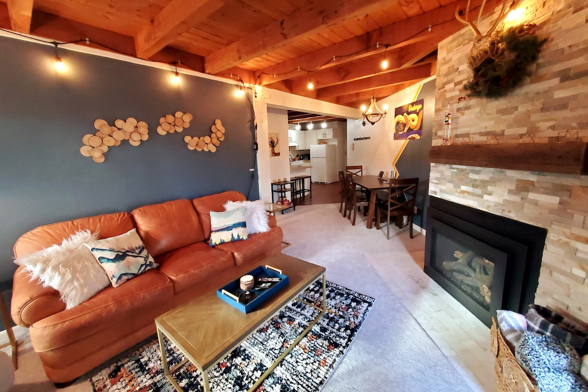 A spacious living area features a leather sofa adorned with decorative pillows. The room is highlighted by a stone fireplace and a rustic wood accent wall. Soft lighting creates a welcoming ambiance, while a dining area can be seen in the background.