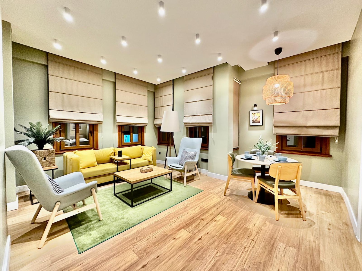 A bright living area is equipped with a yellow sofa and blue accent chairs, arranged around a low coffee table. Soft lighting enhances the warm atmosphere. A dining table with wooden chairs occupies one corner, while large windows feature elegant blinds that filter natural light.