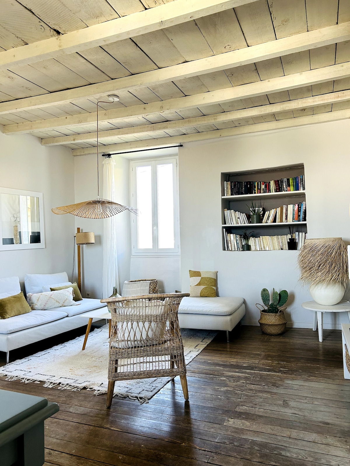 The living area features a modern, light-colored sectional sofa adorned with decorative cushions. A wooden coffee table complements a woven chair, while a bookcase filled with books adds a cozy touch. A large window allows natural light to brighten the space.