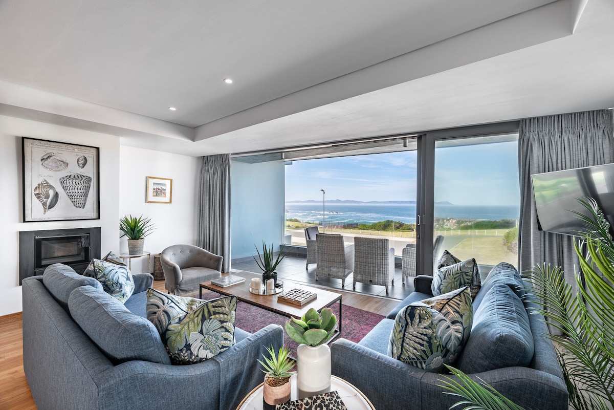 The lounge area features comfortable seating with ocean views through large glass doors. An indoor fireplace is present, and a coffee table is adorned with decorative plants. Natural light fills the space, complemented by neutral-toned furnishings and coastal decor.