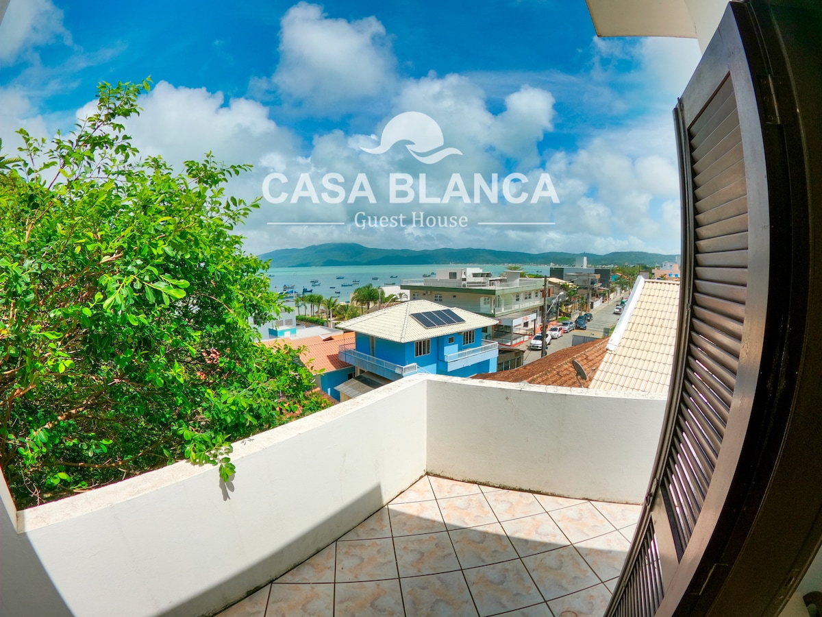 A balcony view showcases a wide expanse of the ocean, framed by lush greenery and distant rooftops. The sky is bright with soft clouds, and the vibrant colors of nearby buildings are visible along the coastline.