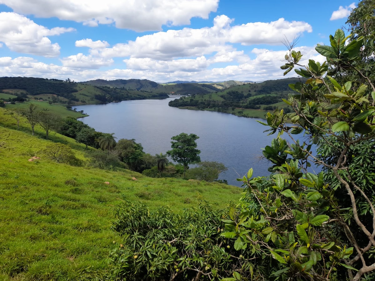 A serene view of a lake is framed by lush green hills under a partly cloudy sky. The tranquil water reflects the surrounding landscape, with trees and grassy banks adding to the natural beauty of the scene.