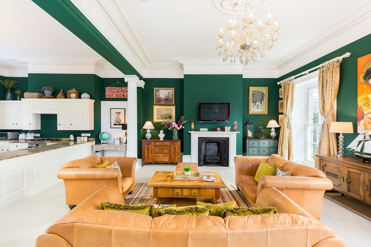 The spacious living area features high ceilings and rich green walls, enhancing its elegance. A chandelier hangs above a central coffee table, surrounded by comfortable leather sofas. Light enters through large windows adorned with drapes, and decorative art pieces add charm to the overall design.