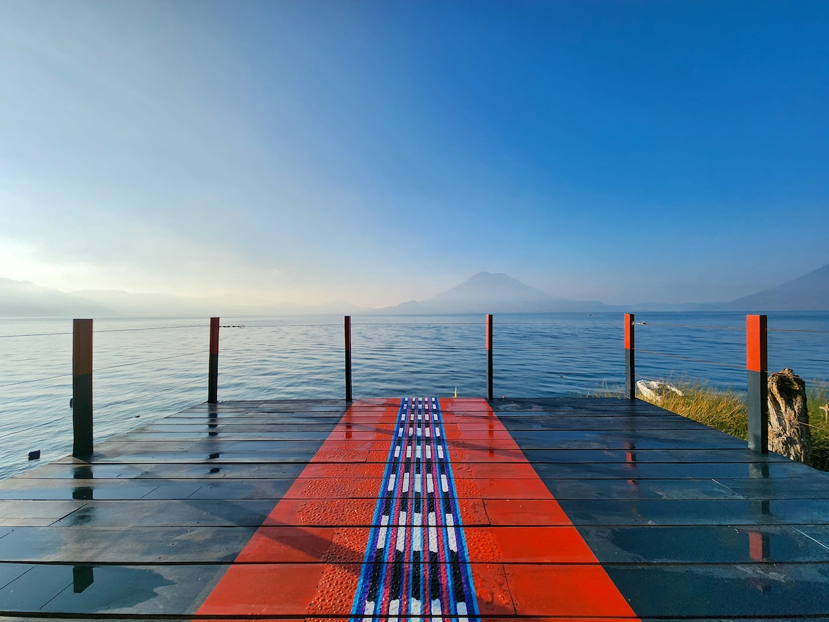 A wooden dock extends over the tranquil waters of Lake Atitlán, featuring bold red accents and intricate patterns along its center. The serene landscape is framed by distant volcanoes under a clear blue sky, creating a peaceful setting for relaxation.