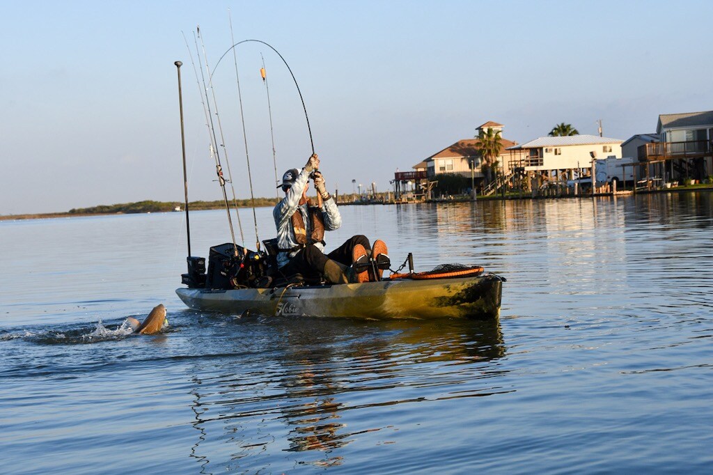 A person is seated in a kayak, actively reeling in a fish while rods are secured in holders. The calm water reflects the surroundings, featuring houses along the canal's edge under a clear blue sky.