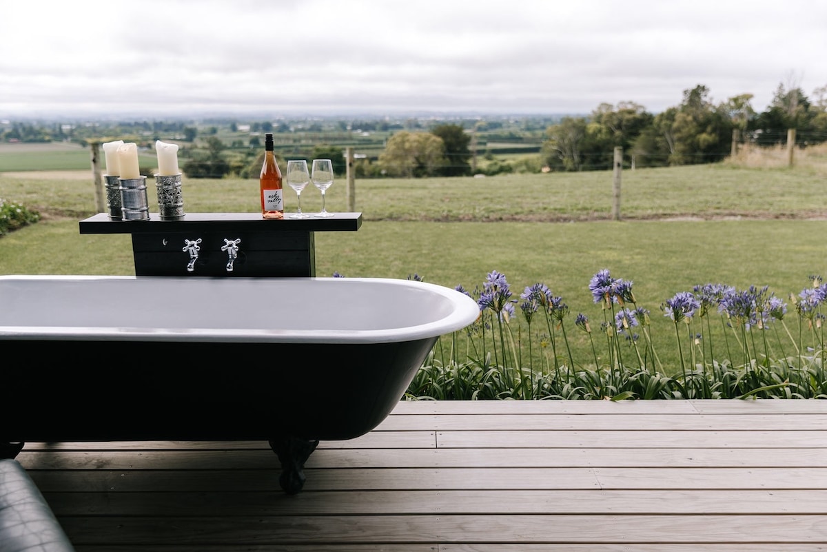 An outdoor bath set on a wooden deck offers a serene view of the expansive green fields and distant hills. A small table nearby displays two glasses and a bottle of wine, complemented by decorative candles and flowering plants lining the edge.