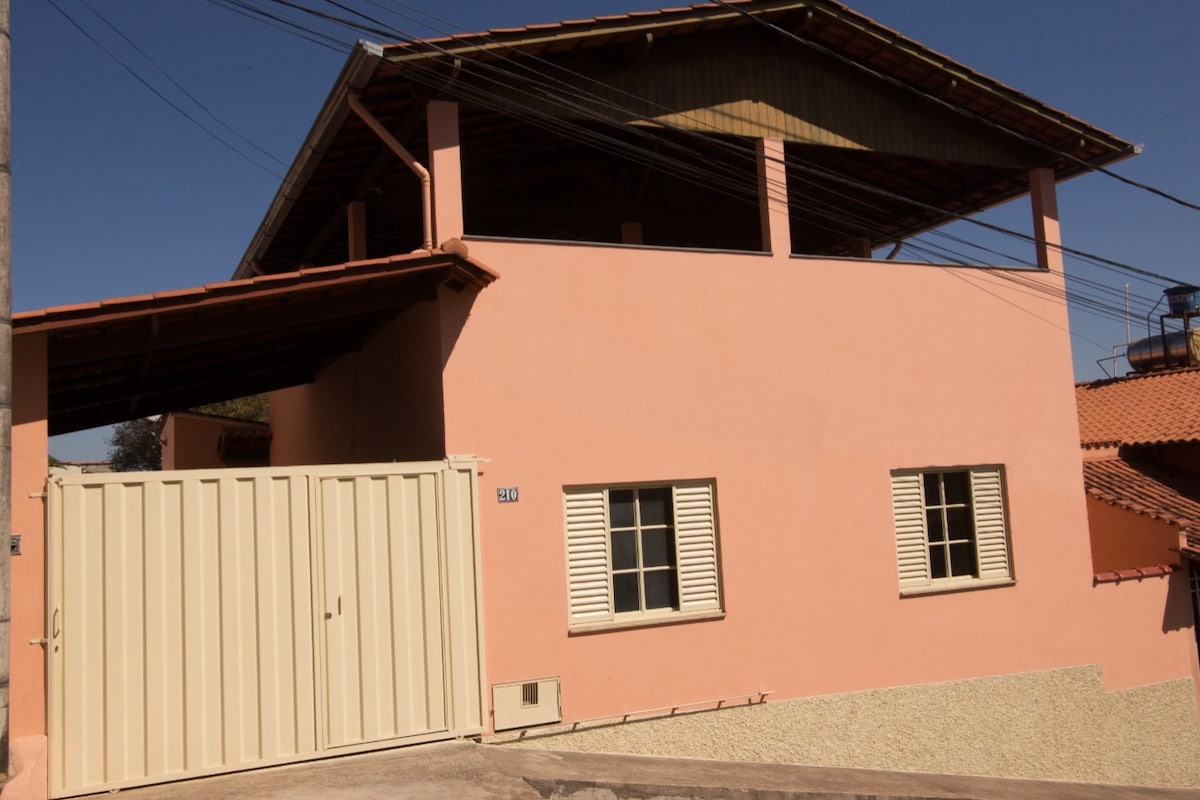 The exterior of a peach-colored house is shown, featuring a distinctive sloped roof and wooden eaves. Secure entry is provided by a metal gate, while window shutters add texture to the façade. Surrounding structures are partially visible, indicating a residential environment.