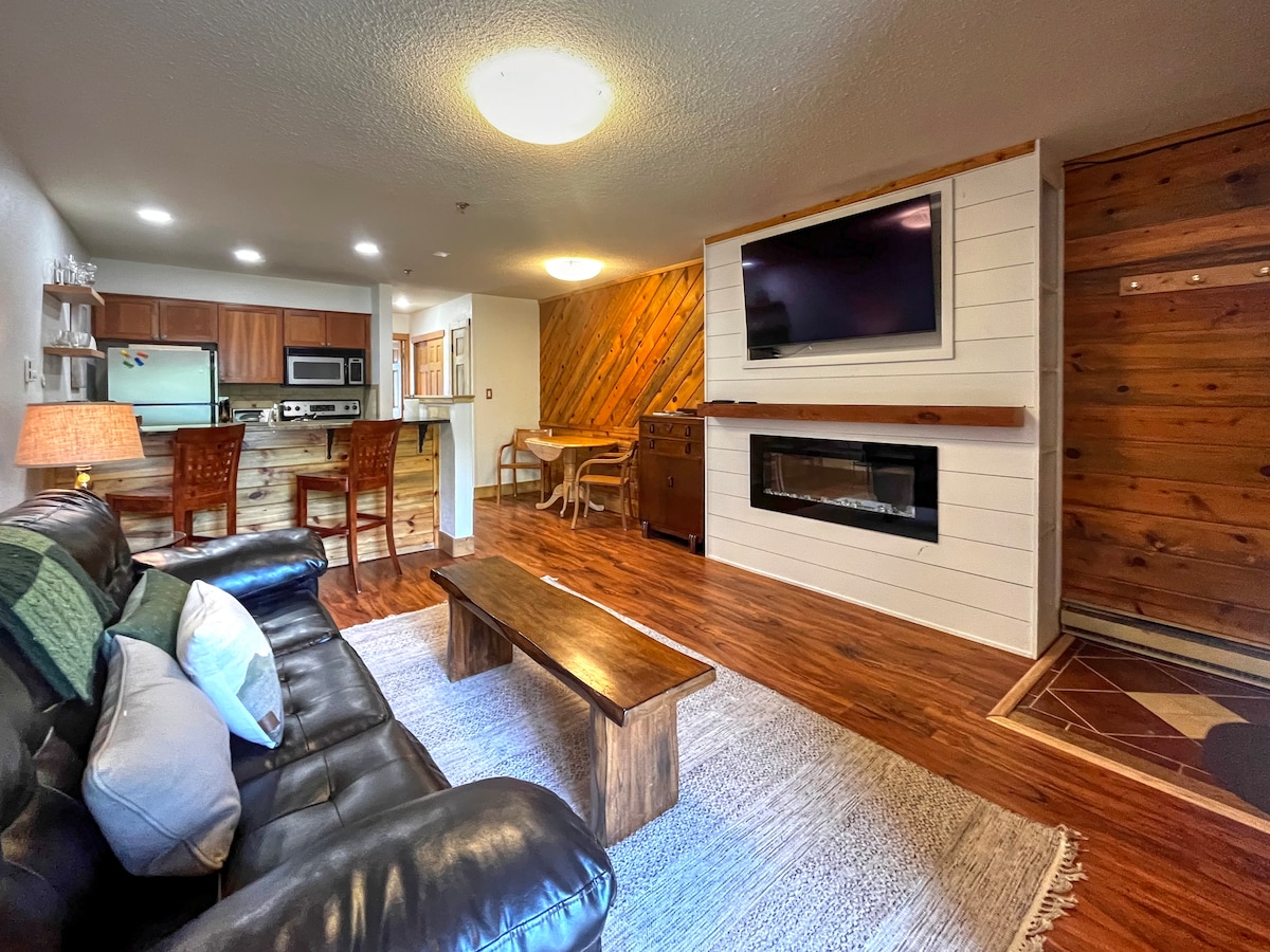 The living area features a dark leather sofa with decorative pillows and a central wooden coffee table. A flat-screen TV is mounted on a white shiplap wall above a modern fireplace. The open layout reveals the kitchen and dining area, showcasing wood accents and warm lighting.