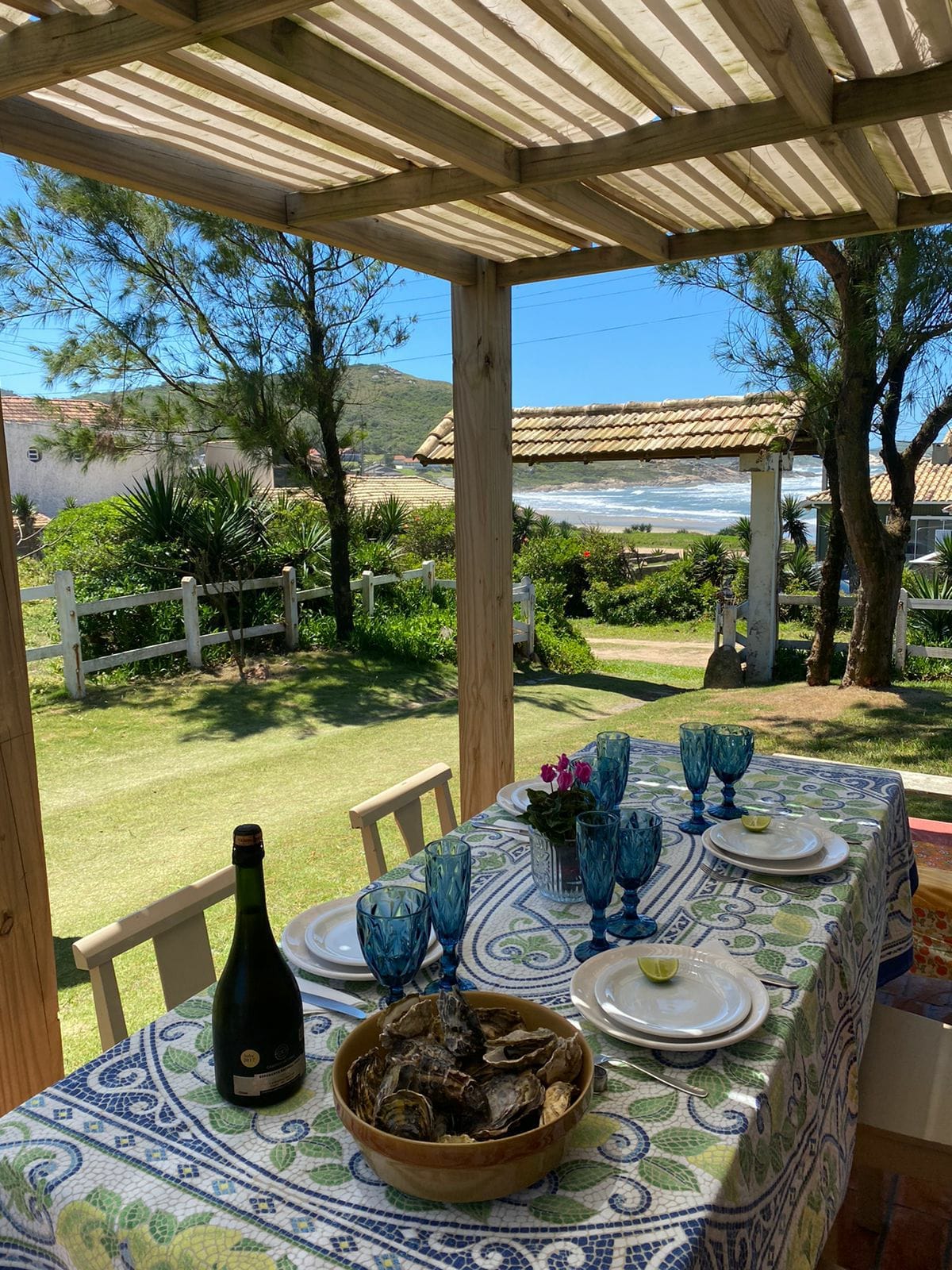 A dining area is set under a shaded outdoor structure, featuring a colorful tablecloth. The table is arranged with plates, glasses, and fresh oysters. A view of the lush garden and the beach can be seen in the background, illuminated by sunlight.
