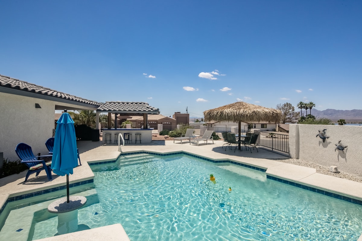 A private pool with crystal blue water reflects the sky, surrounded by a patio featuring lounge chairs and a shaded Tiki Hut. A bright blue umbrella adds a pop of color, while palm trees and distant mountains create a serene backdrop.