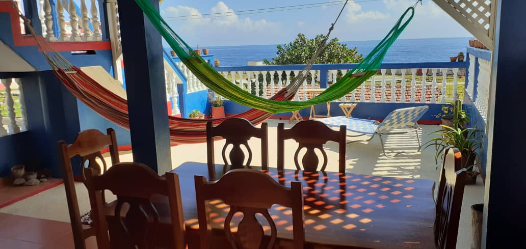 An outdoor terrace is presented with a large wooden dining table accompanied by several chairs. Hammocks in vibrant colors are suspended in the background, overlooking a serene ocean view. Lush greenery is visible, along with a sun loungers positioned for relaxation.