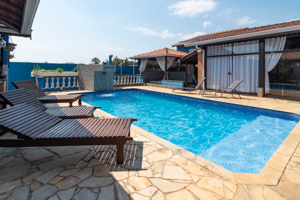 A spacious outdoor pool area features a large swimming pool with crystal-clear water. Sun loungers are arranged along the edge, offering a place to relax. The surrounding patio is made of natural stone, and a covered area with seating is visible in the background.