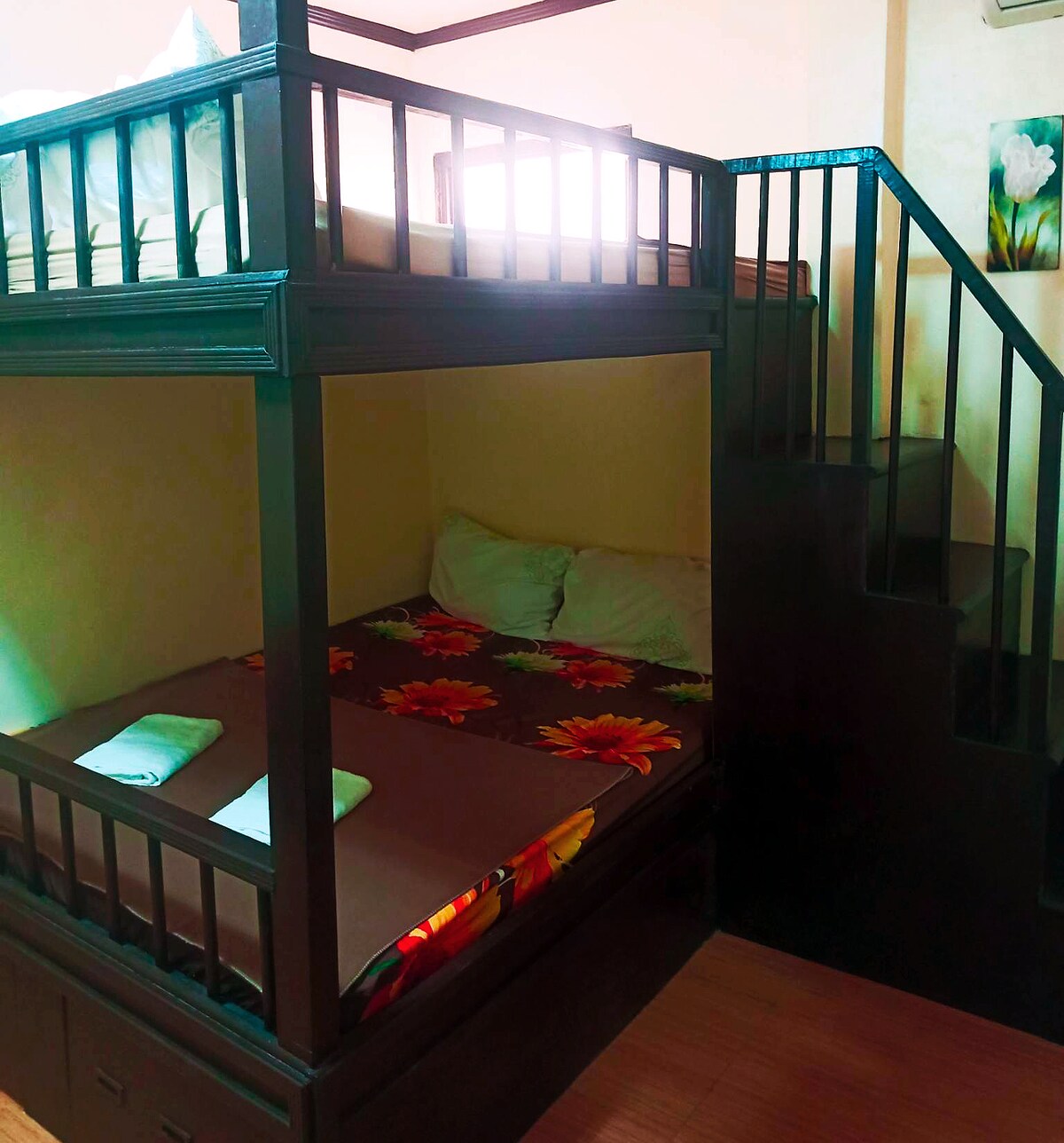A multi-level bunk bed is featured, with a spacious lower sleeping area adorned with floral-patterned bedding and two white pillows. The upper level includes a neatly made bed with light-colored linens. Stairs lead to the upper bunk, with natural light entering from a nearby window.