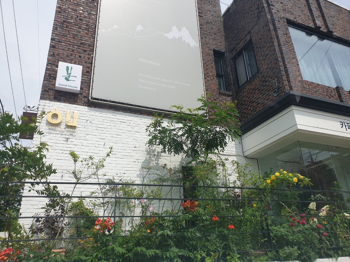 The exterior of a building is showcased, featuring a mix of brick and white walls. A garden with various flowers and greenery adds a natural element to the scene, providing a welcoming entrance. A sign with the word 'Himalaya' is prominently displayed.
