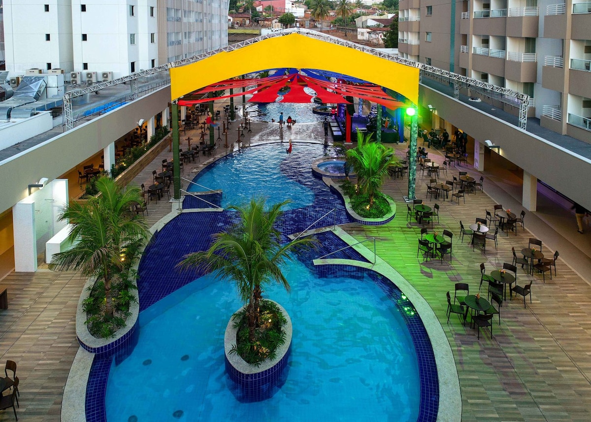 An expansive outdoor pool area is surrounded by palm trees and lounge chairs. Colorful canopies provide shade, creating a lively atmosphere. The pool features various sections, with clear blue water reflecting the surrounding lights, inviting relaxation and leisure.