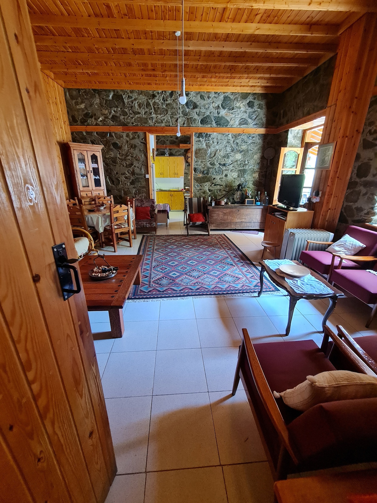 A cozy living area features natural stone walls and wooden accents. Comfortable seating is arranged around a central area rug, with a dining space visible in the background. Sunlight filters through large windows, enhancing the welcoming atmosphere of the room.