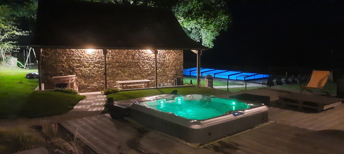 An outdoor area is illuminated by soft lighting at night, featuring a hot tub with cascading water and a serene green glow. The covered outdoor structure sits adjacent to a pool with a protective cover, surrounded by a spacious wooden deck for relaxation.