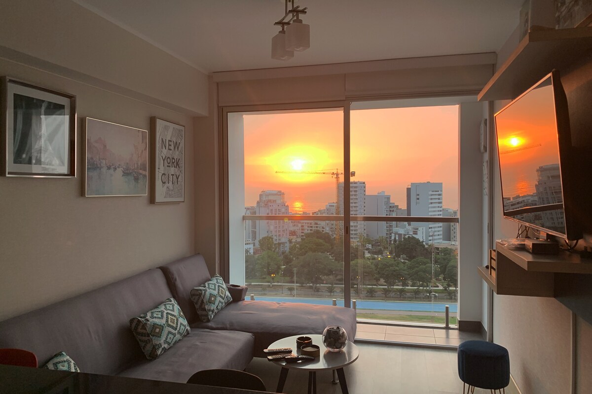 A living area features a comfortable gray sofa adorned with decorative pillows. Large sliding glass doors provide a stunning sunset view over the city skyline. A flat-screen television is mounted on the wall, and a small round table is positioned nearby.