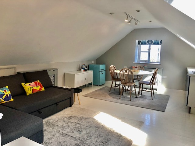 A contemporary living area features a dark sofa adorned with colorful cushions, a light rug beneath, and wooden dining chairs surrounding a round table. A kitchenette is visible, coupled with large windows allowing natural light to fill the space, enhancing the openness.