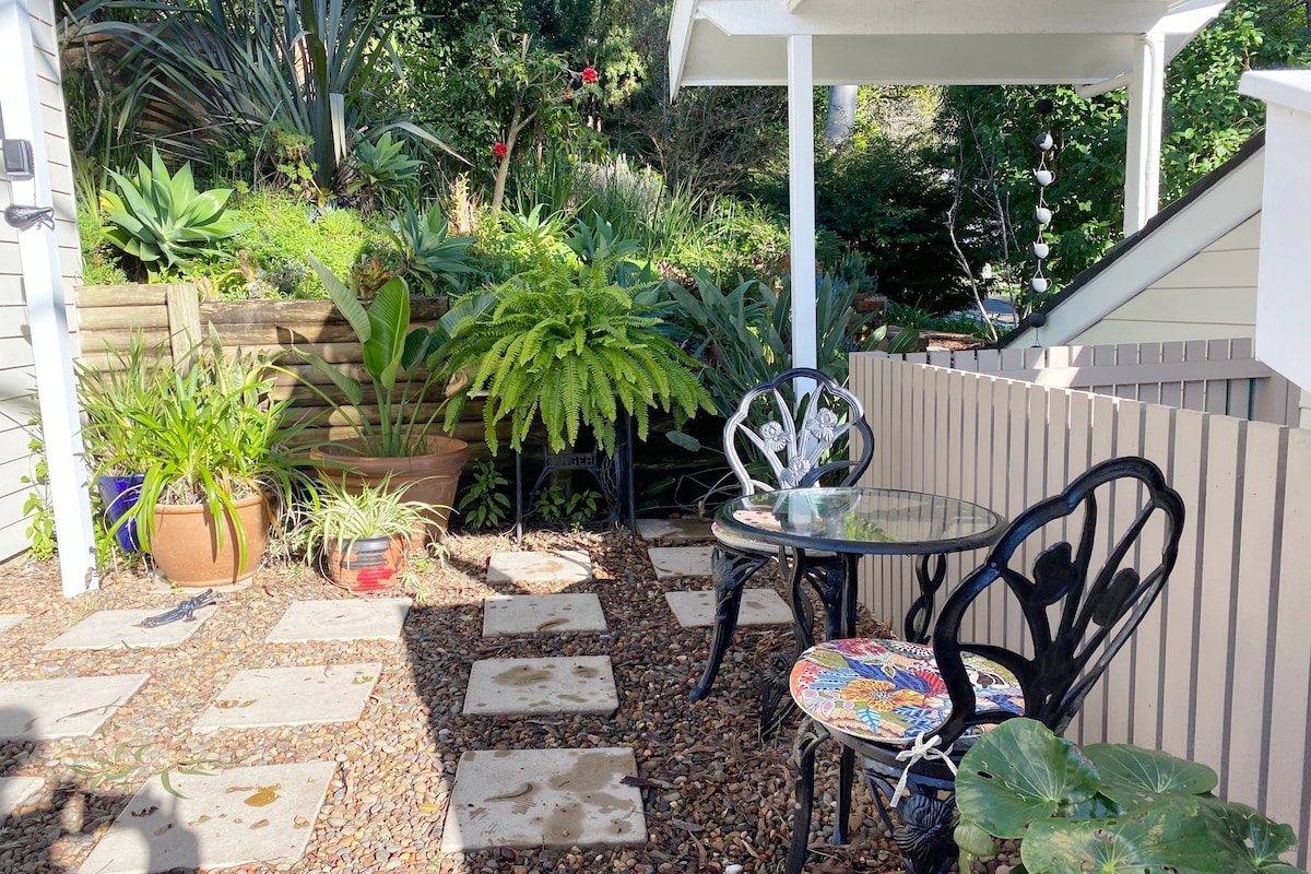 An outdoor courtyard is framed by lush greenery, featuring two black chairs with decorative cushions around a glass table. Stone pavers create a pathway, while potted plants add vibrant colors to the serene space.