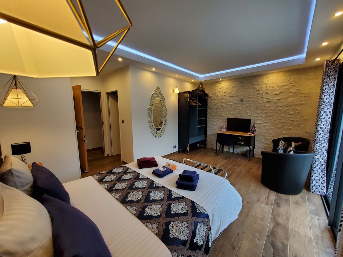 A spacious bedroom features a queen-size bed adorned with layered linens and decorative pillows. Modern furnishings include a wardrobe and a desk, while a flat-screen TV is positioned on a wall. Natural light floods in from floor-to-ceiling windows, enhancing the neutral tones of the room.