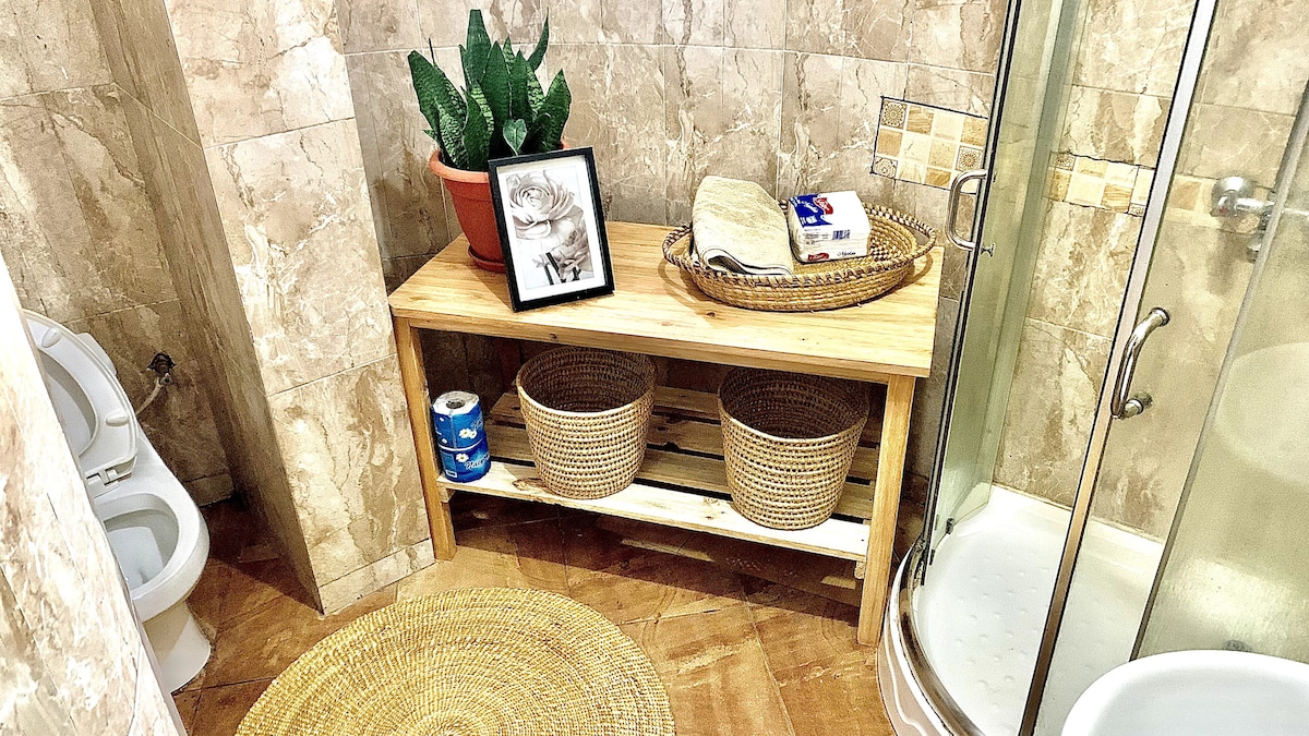 A well-appointed bathroom features a clean, modern shower and a toilet. A wooden bench holds decorative items, including a framed picture, a plant, and essential toiletries. Two woven baskets provide additional storage, while a round rug adds warmth to the tiled floor.