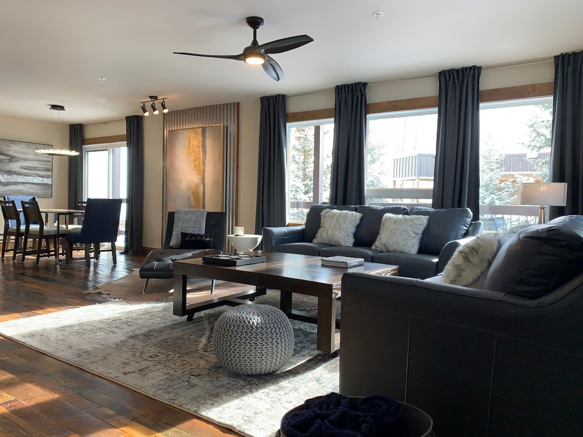 A spacious living area features two comfortable sofas and a decorative pouf, surrounded by large windows that allow natural light to fill the space. Dark curtains frame the windows, and a dining table is seen in the background, adding to the inviting atmosphere.