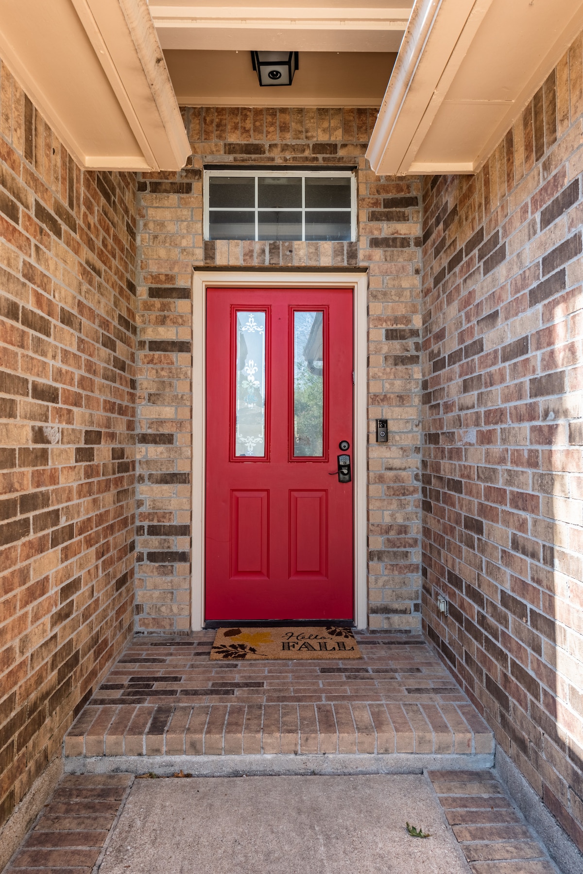 A warmly colored red door welcomes guests to the entryway, framed by a stylish brick exterior. A welcome mat is placed on the tiled step, adding a friendly touch. Natural light filters in through the top window above the door.